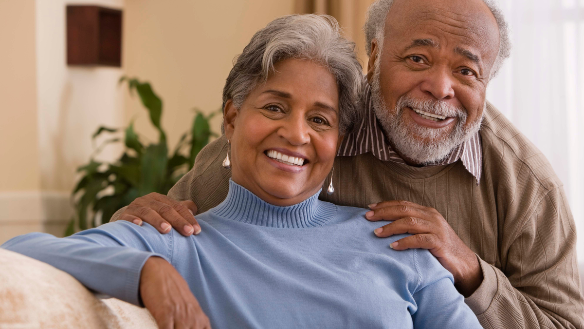 An elderly African American couple smiling warmly in a cozy indoor setting. The woman in the foreground wears a light blue turtleneck sweater, while the man behind her wears a brown jacket over a striped shirt.